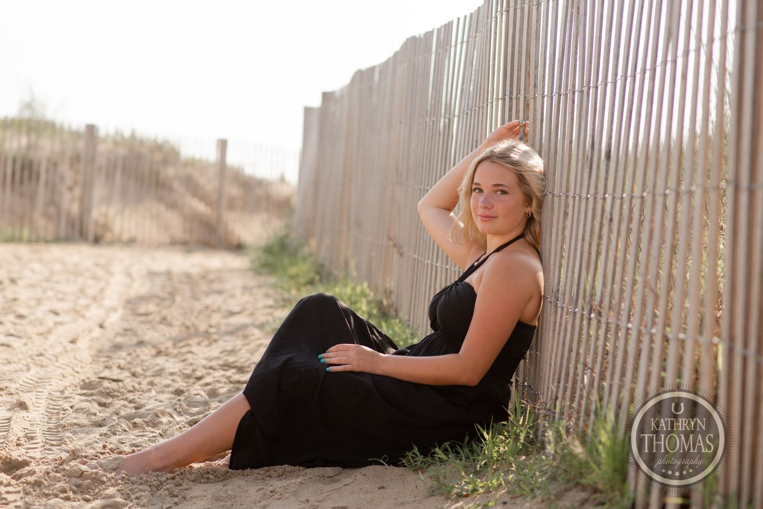 Natural light portrait by beach fence wearing black sundress.