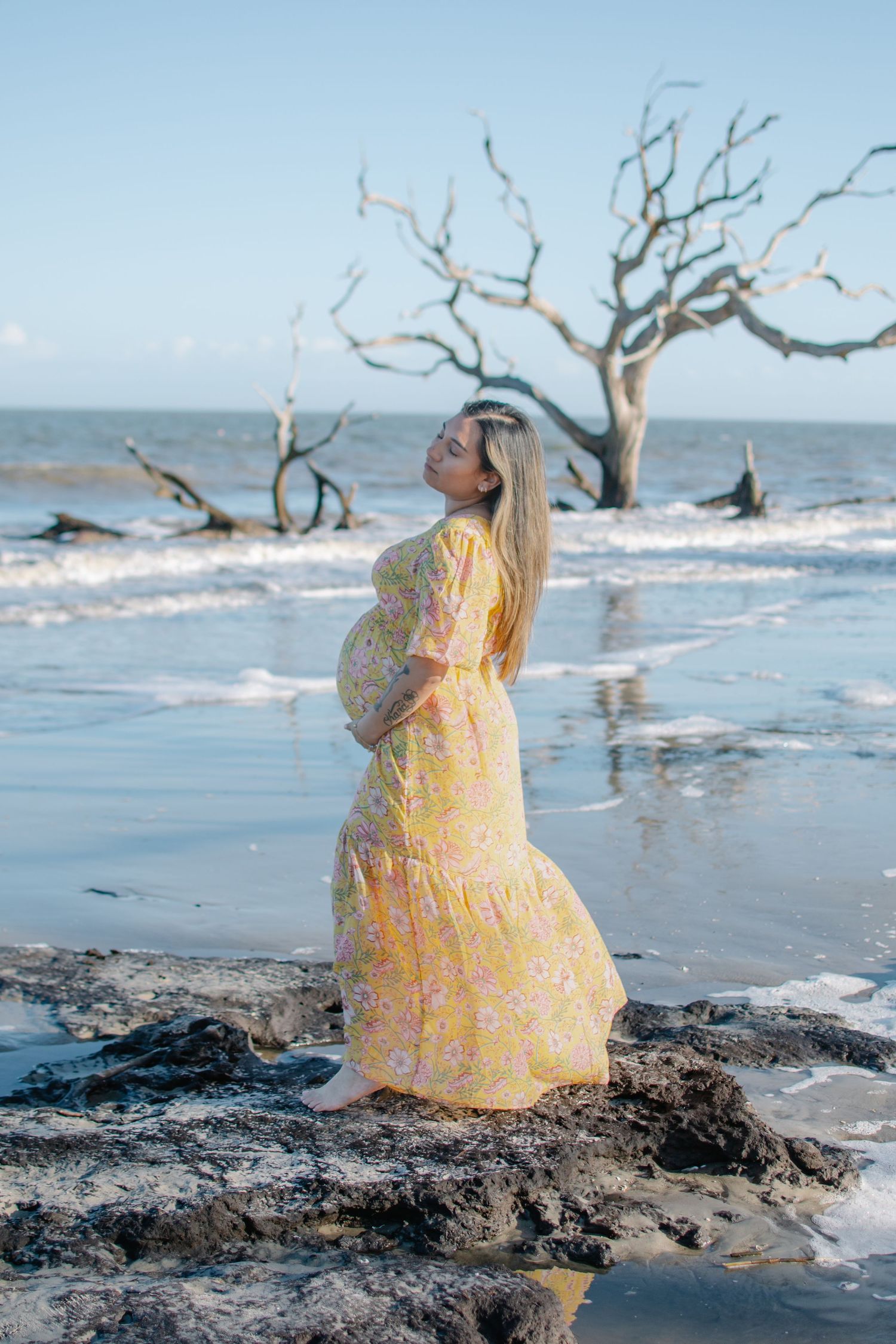 Expectant figure in yellow dress poses on beach with driftwood trees at sunset.
