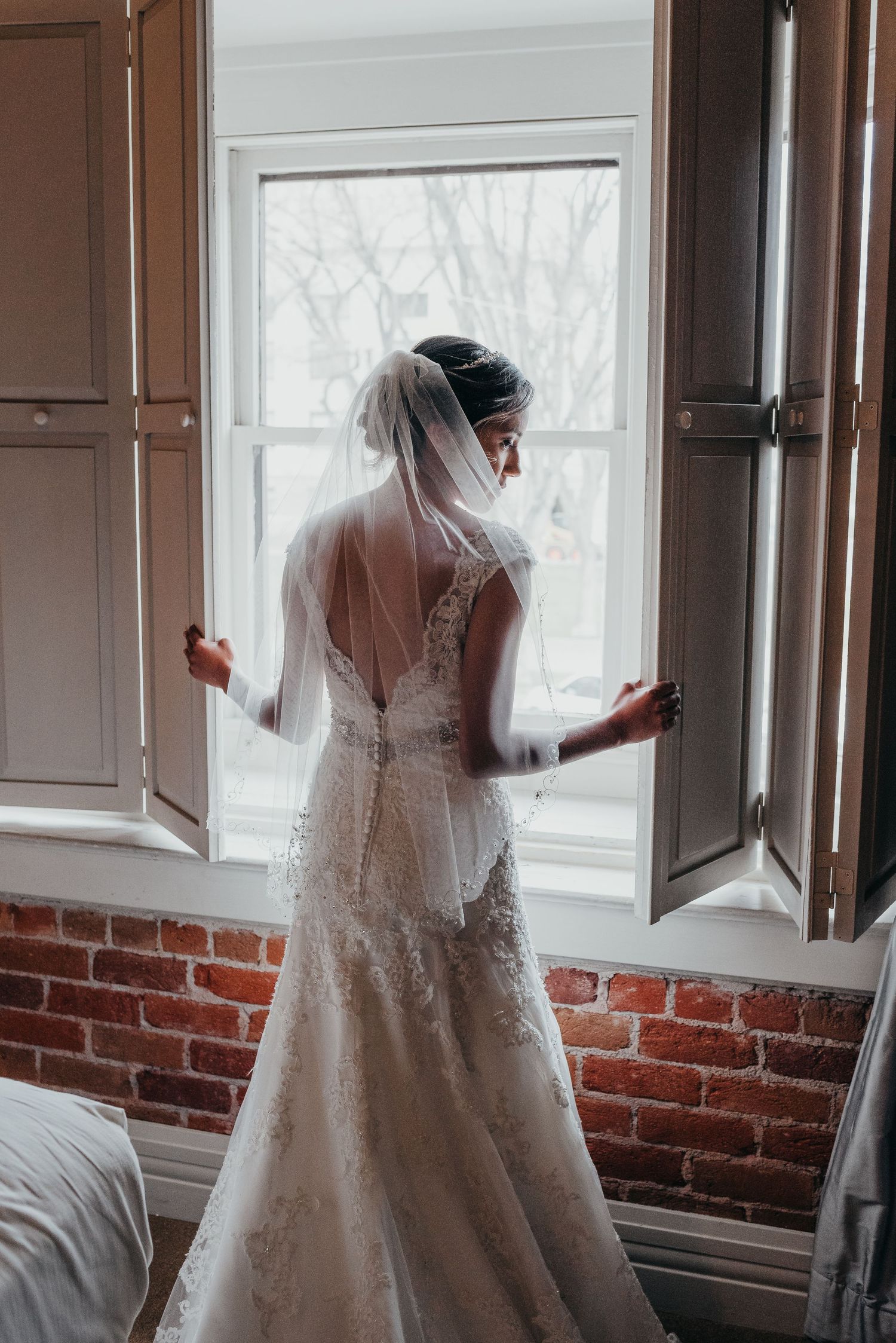 A bride in a lace wedding dress with an open back stands at a window with exposed brick walls.