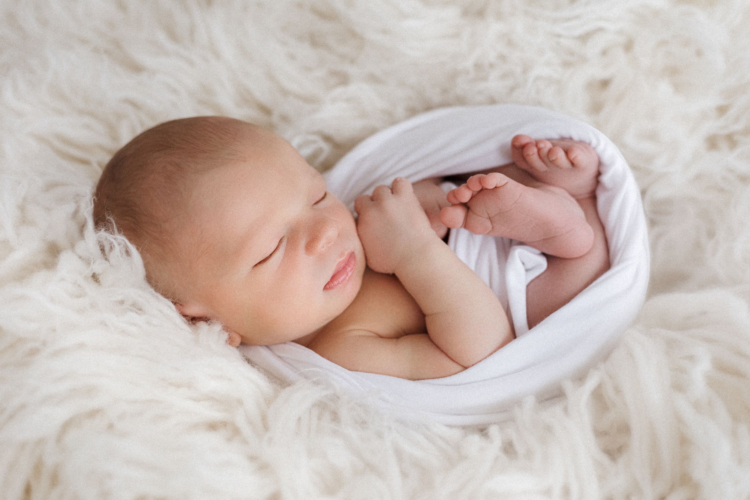 Sleeping newborn wrapped in white fabric on fluffy cream-colored blanket.