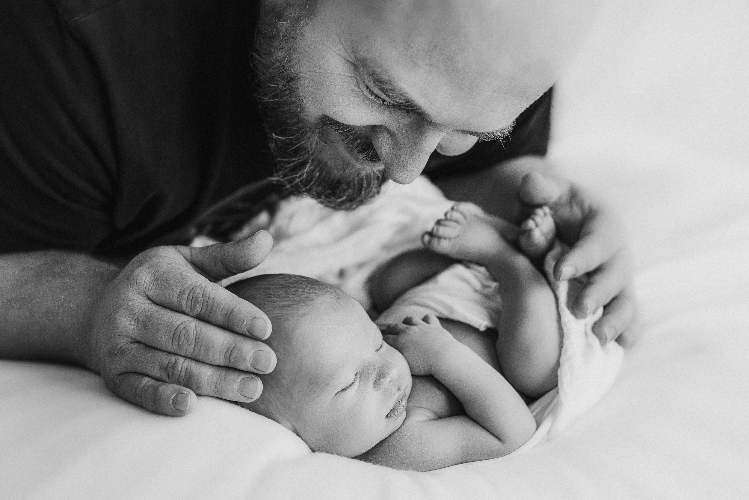 A tender black and white portrait of a newborn baby being cradled against soft white bedding.