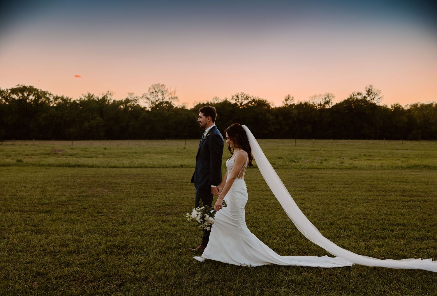 Newlywed couple with flowing veil walking in grassy field at sunset.