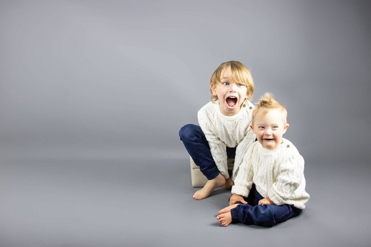sibling giggling while having there photo taken In photography studio wearing matching jumpers and jeans, gray back drop
