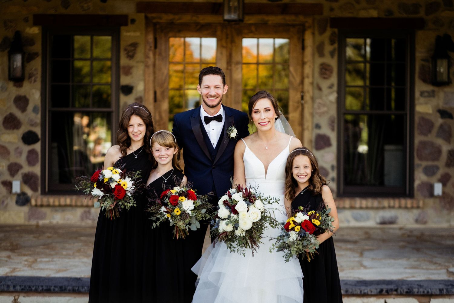 Wedding party in formal attire posed in front of a rustic stone building at sunset.