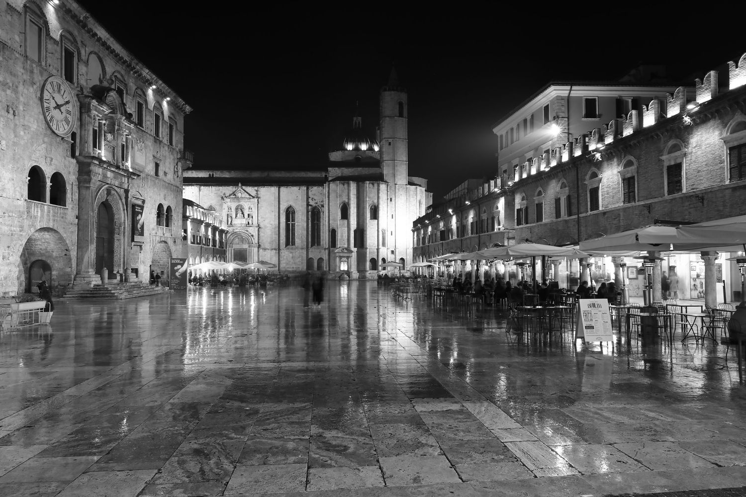 Rainy night scene of Ascoli Piceno's main square with historic buildings reflected in wet pavement.