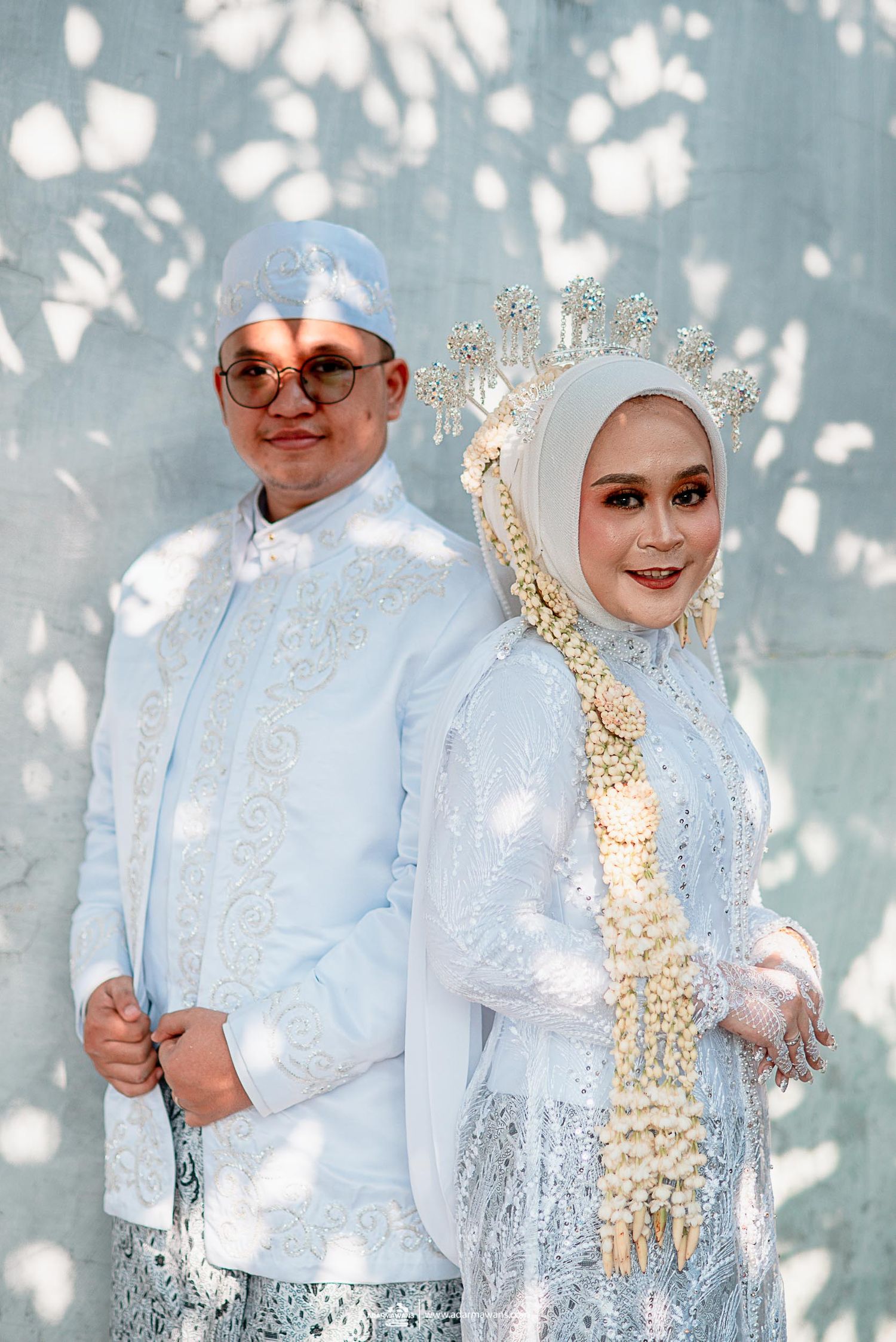 Muslim wedding couple in matching light blue traditional attire against dappled light background.