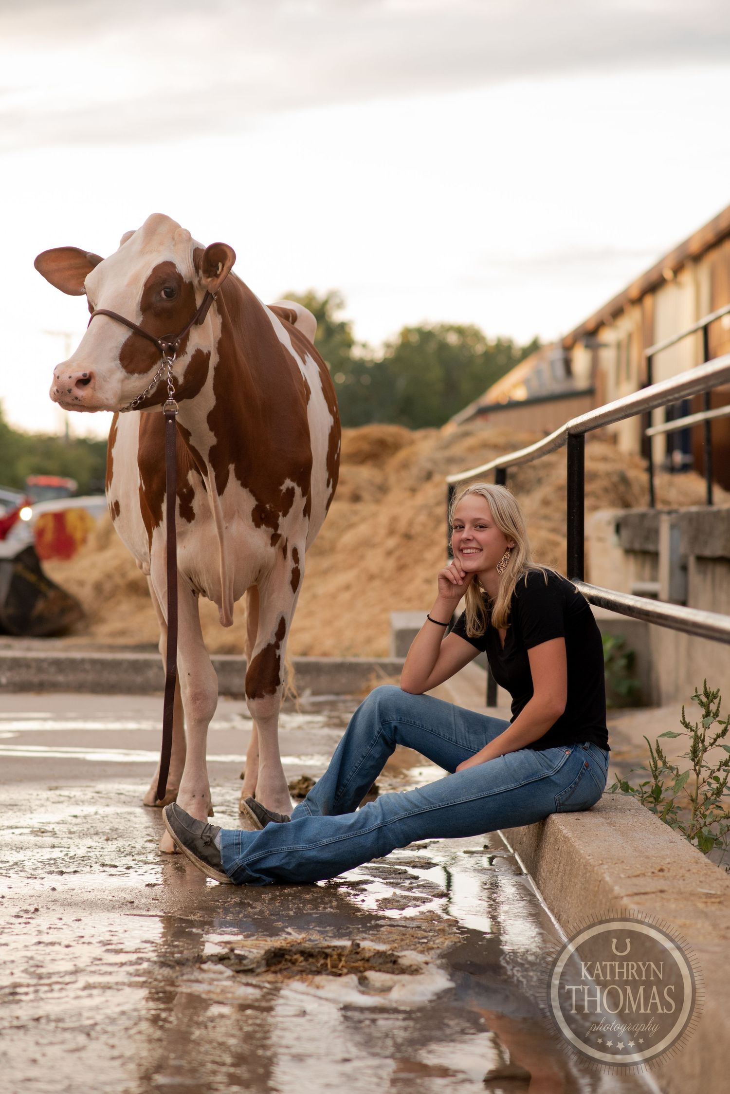 A Holstein dairy cow stands near a person sitting on concrete steps with farm buildings in background.