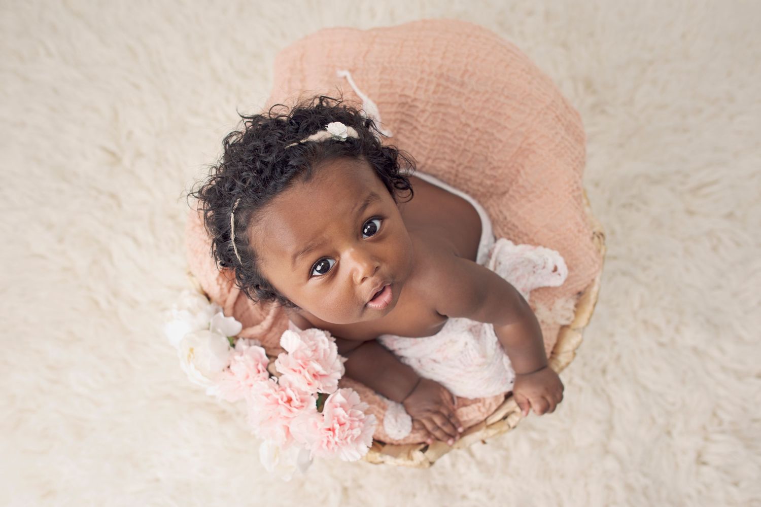 A baby poses in a round basket with pink flowers during an elegant studio portrait session.