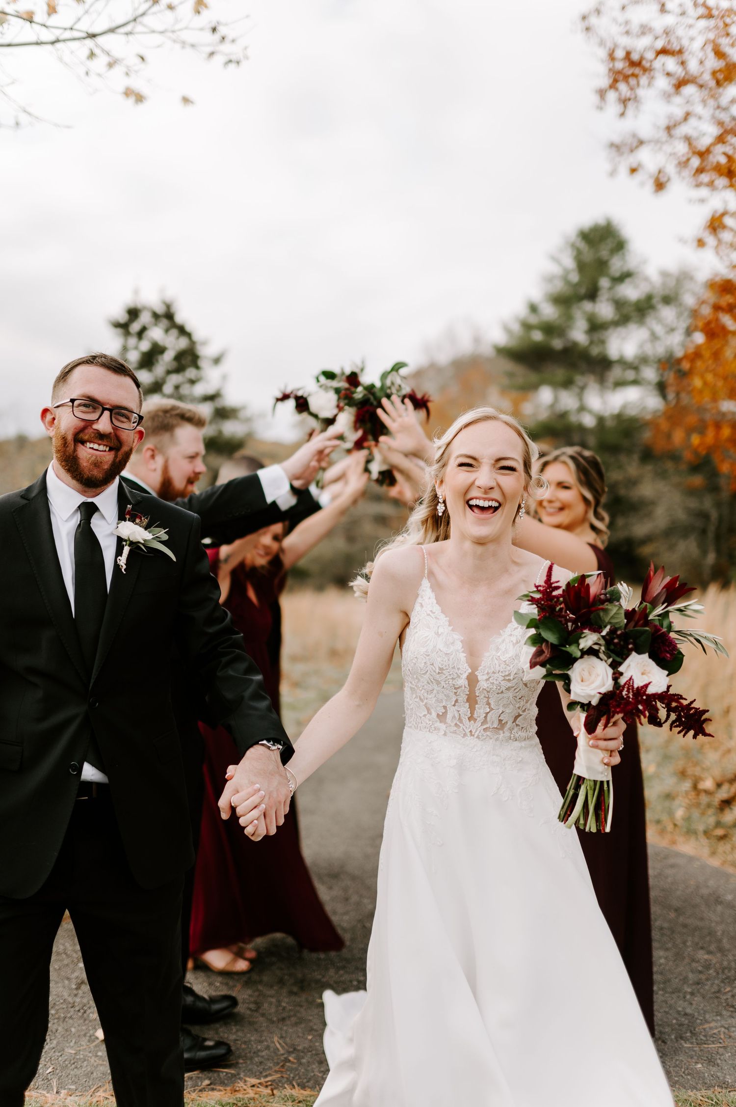 Wedding couple shares joyful moment with bridal party in burgundy dresses against autumn foliage backdrop.