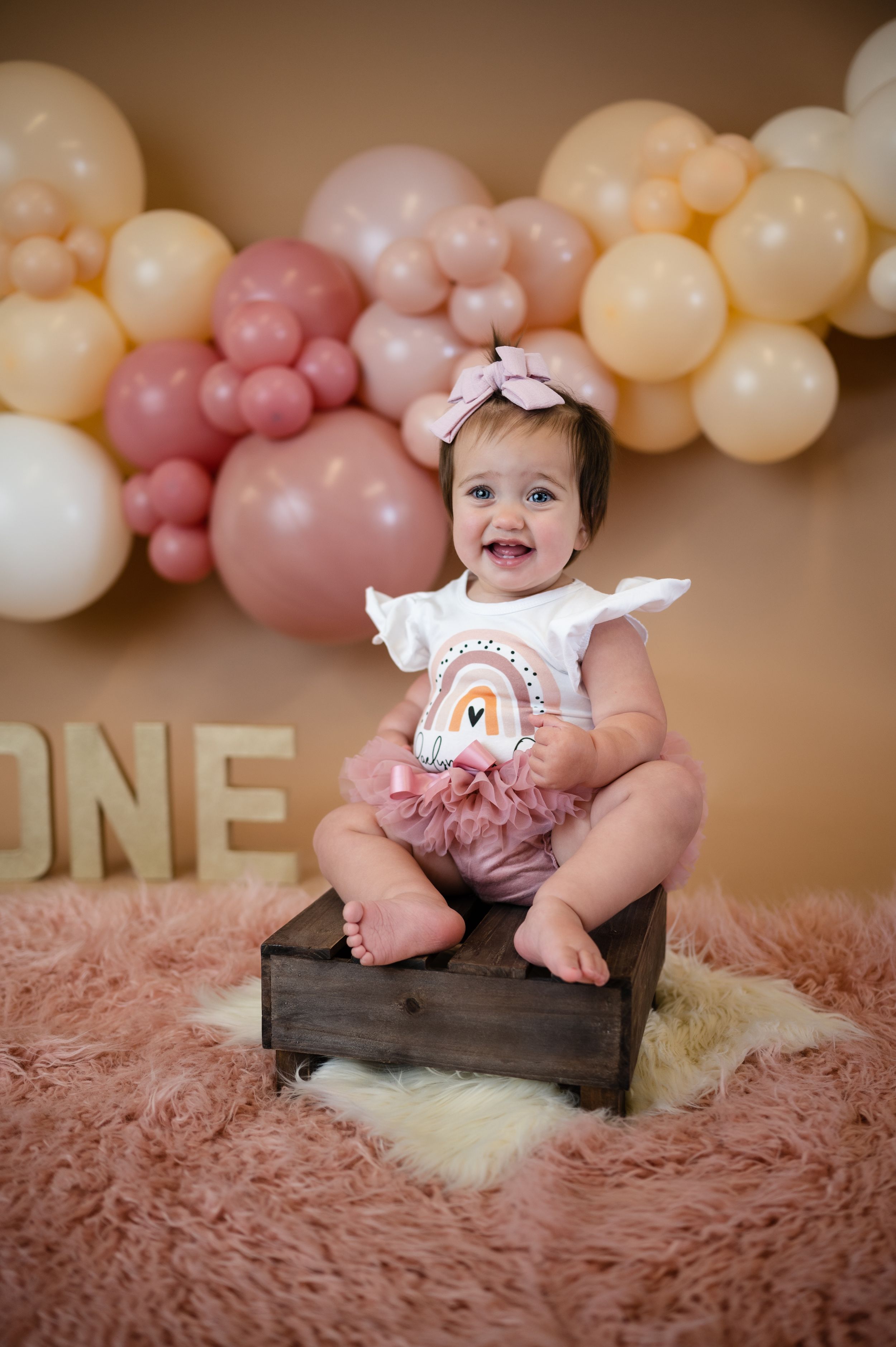 Baby sitting on wooden box with balloon arch backdrop and 'ONE' display for first birthday.