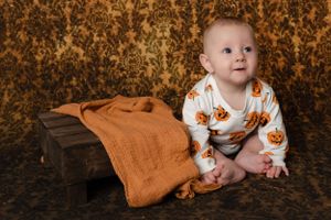 Baby wearing pumpkin print outfit sitting next to wooden prop and orange blanket.