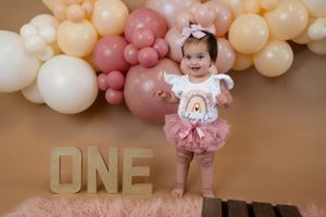First birthday photo with pink and cream balloon arch, gold 'ONE' letters, and pink plush rug.