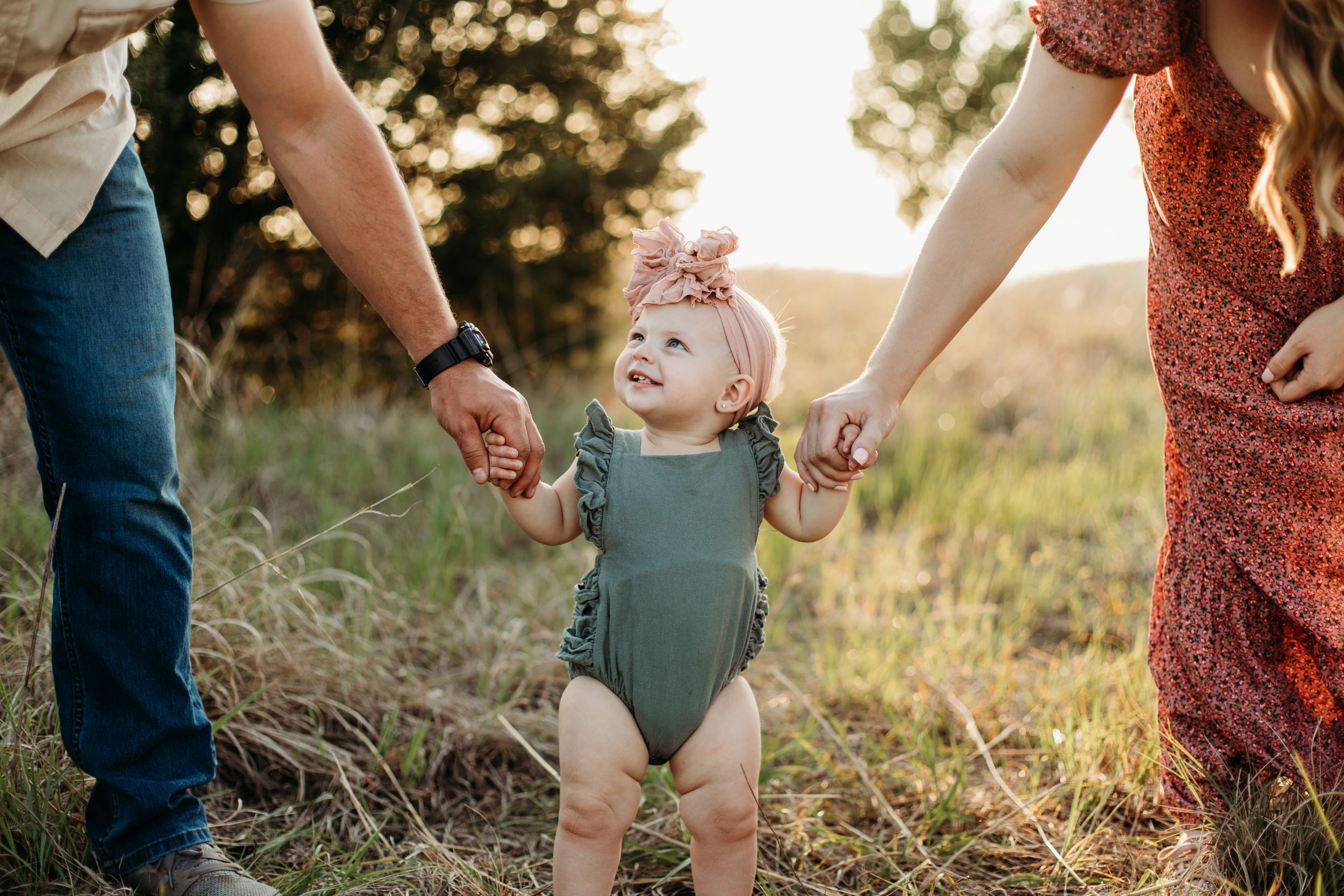 Two adults hold hands with little one in sage romper during golden hour outdoor photoshoot.