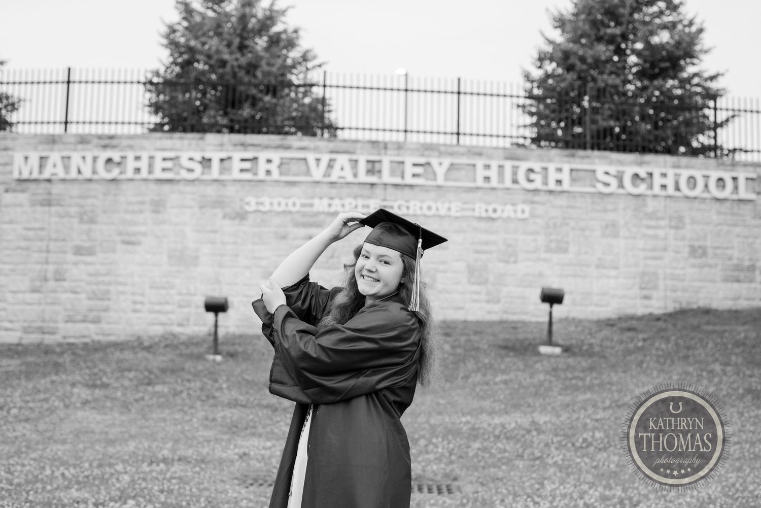 Black and white portrait of graduate posing joyfully in cap and gown outdoors.