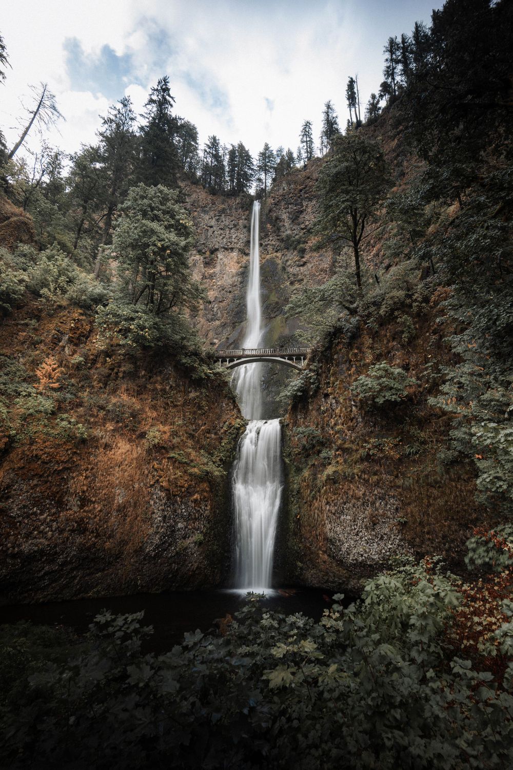 Majestic Multnomah Falls cascades down moss-covered cliffs in Oregon's Columbia River Gorge on a cloudy day.