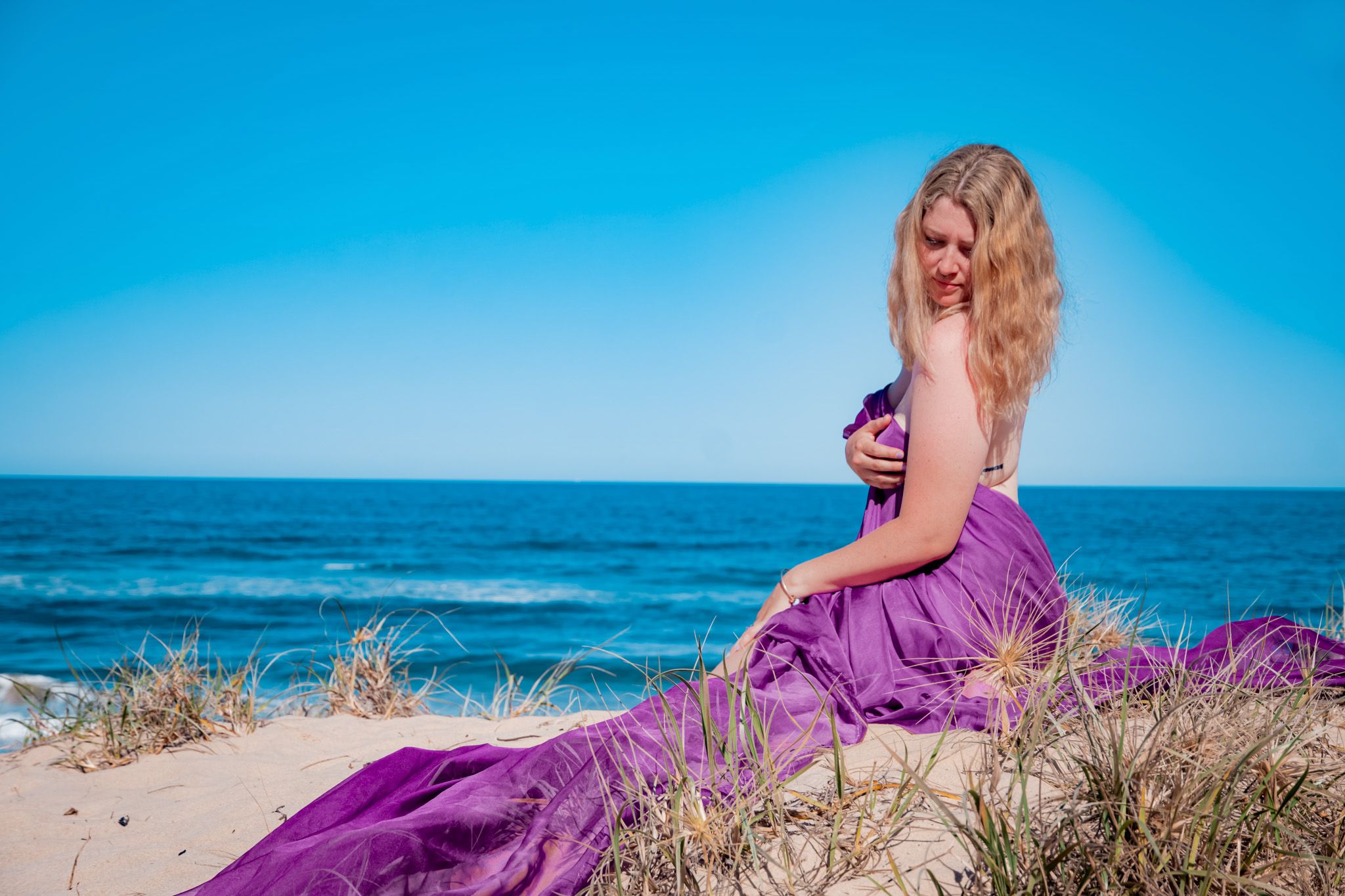 Woman in flowing purple dress posing on beach with ocean backdrop.