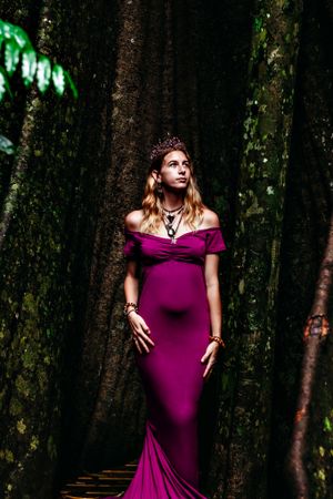 Maternity photoshoot in purple gown against dark forest backdrop with green lighting.