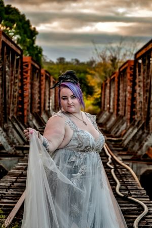 Dramatic portrait in silver gown against rustic railroad tracks at dusk.