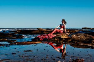 Coastal photoshoot at rocky beach tide pools with flowing red dress against blue sky.