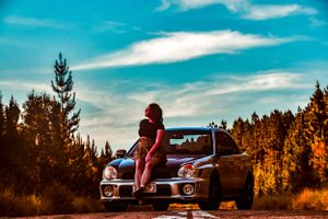 Classic car parked on rural road during golden hour with pine forest backdrop.