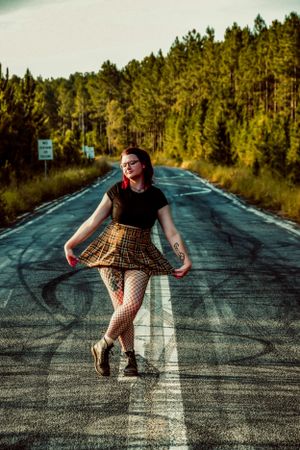 Person in dress playfully posing on an empty road lined with pine trees.