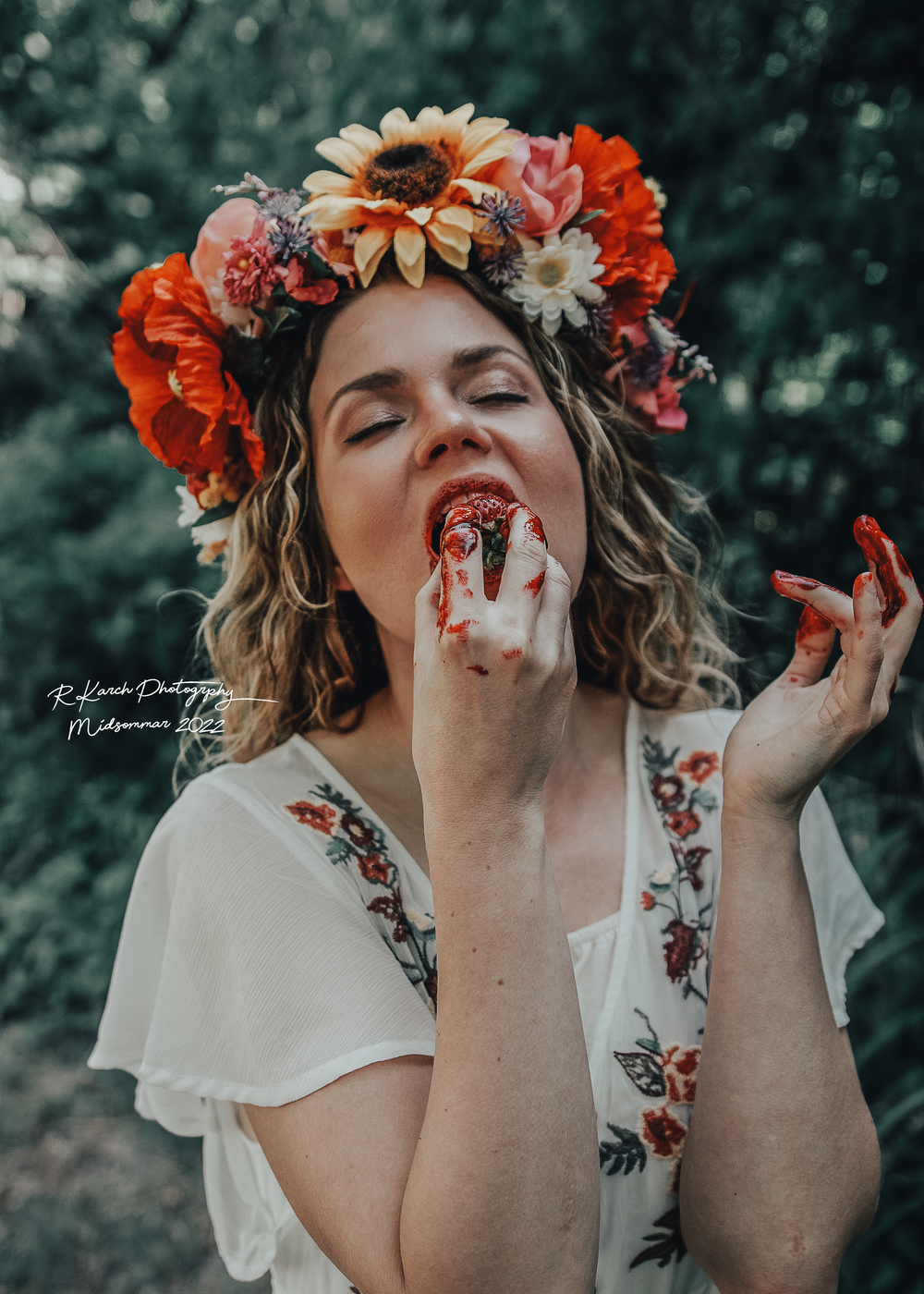 Artistic portrait showing strawberry-stained fingers and vibrant floral crown in dreamy forest setting.