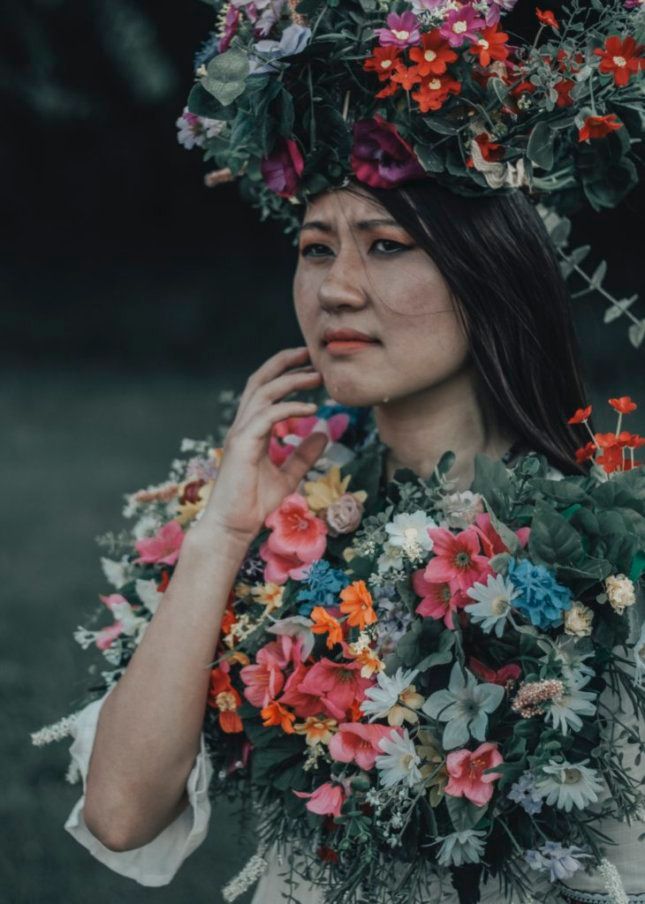 Person wearing floral crown and surrounded by colorful blooms in moody forest setting.