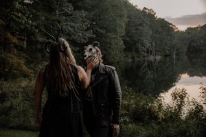 Romantic forest photoshoot at sunset with two figures in dark attire and masks by a lake.