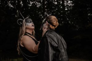 Gothic-styled couple in masks and dark clothing pose dramatically against forest backdrop.