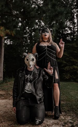 Gothic forest scene with two figures in leather, masks and dark attire striking dramatic poses.