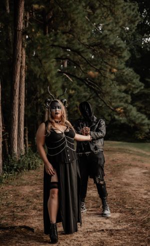Dark fantasy photoshoot in a forest with two figures in gothic attire and masks creating a dramatic atmosphere.