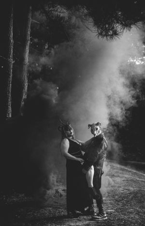 Moody black and white photo of two masked figures embracing in misty forest setting.