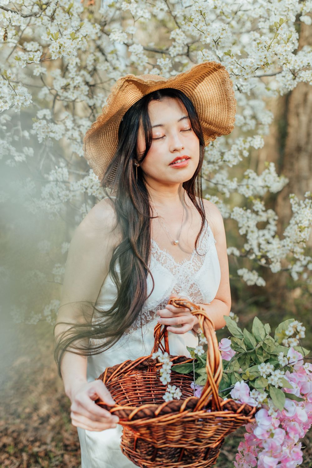 Dreamy spring portrait with woven basket and cherry blossoms featuring a woman in white dress and straw hat.