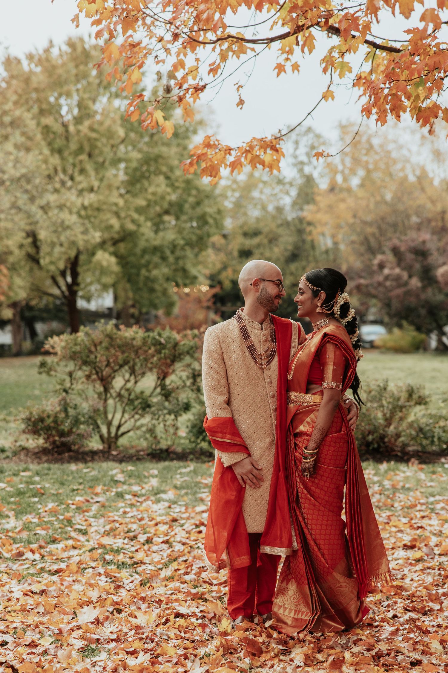 A couple in traditional Indian wedding attire poses among fallen autumn leaves.