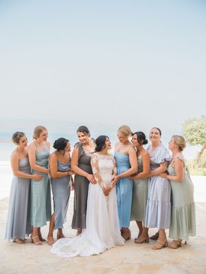 A bridal party in mixed blue and grey dresses poses together on a sunny beach.