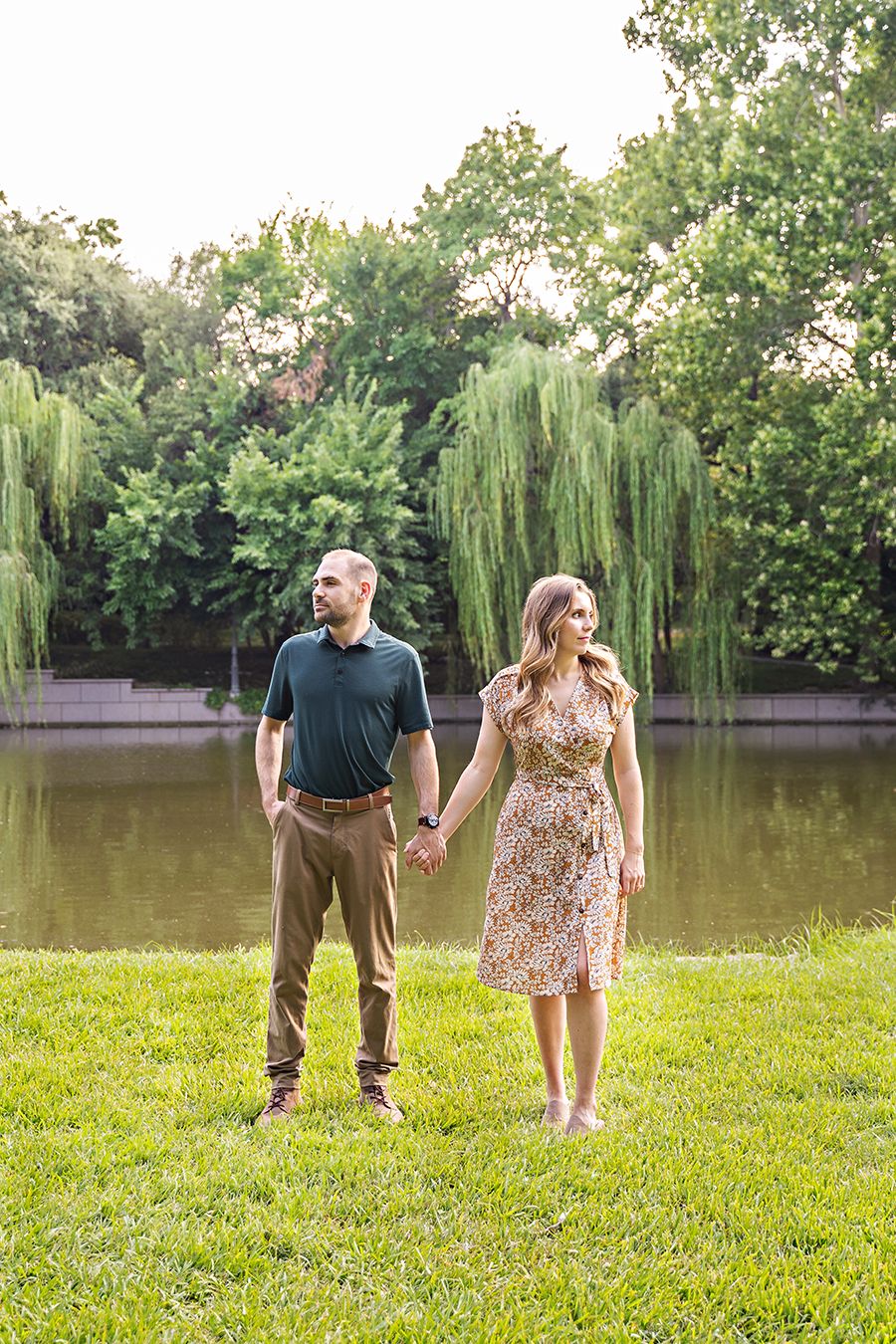 Couple in casual attire enjoys a playful moment by a pond with weeping willows.