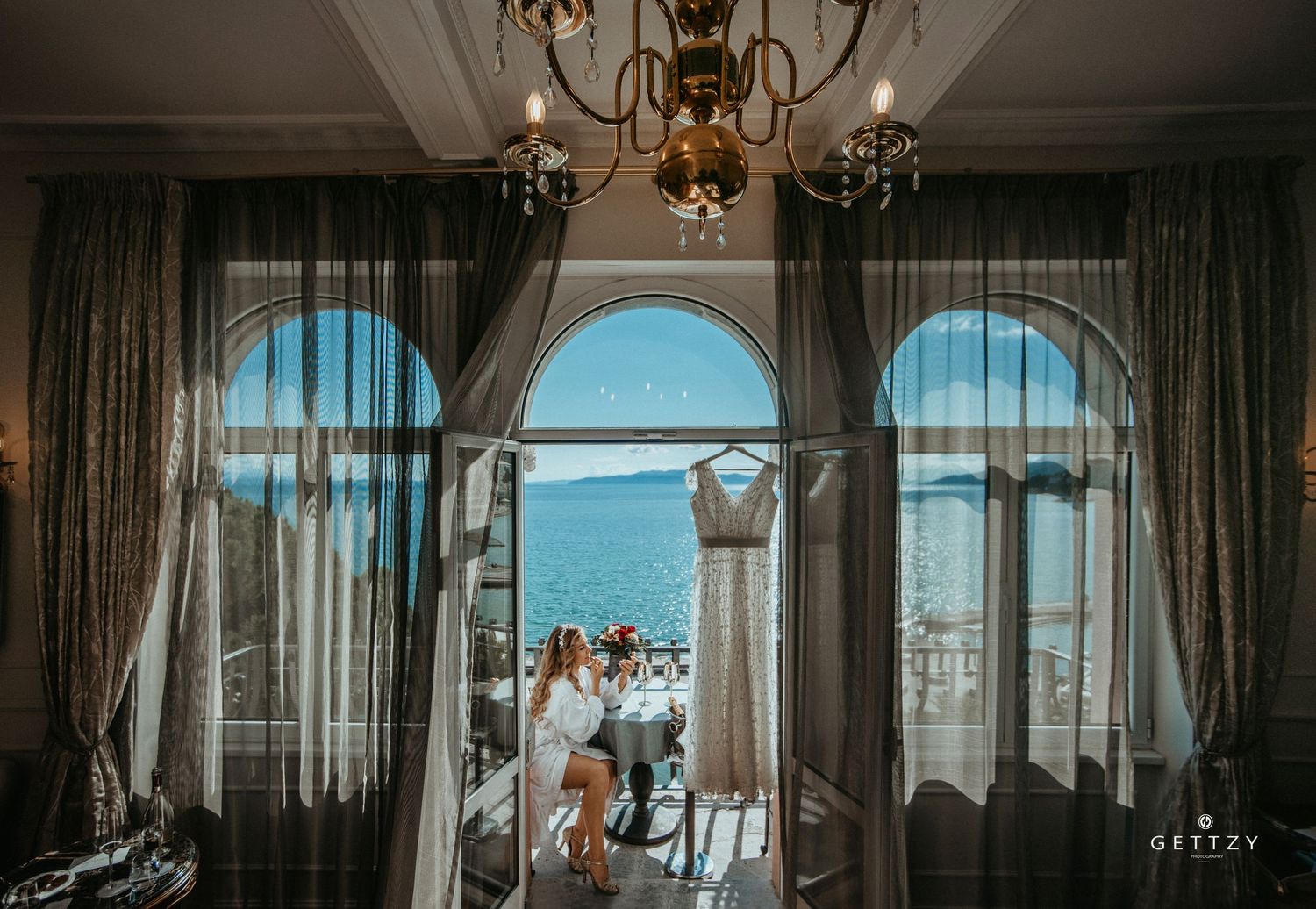 Elegant wedding scene through arched windows overlooking ocean view with chandelier.