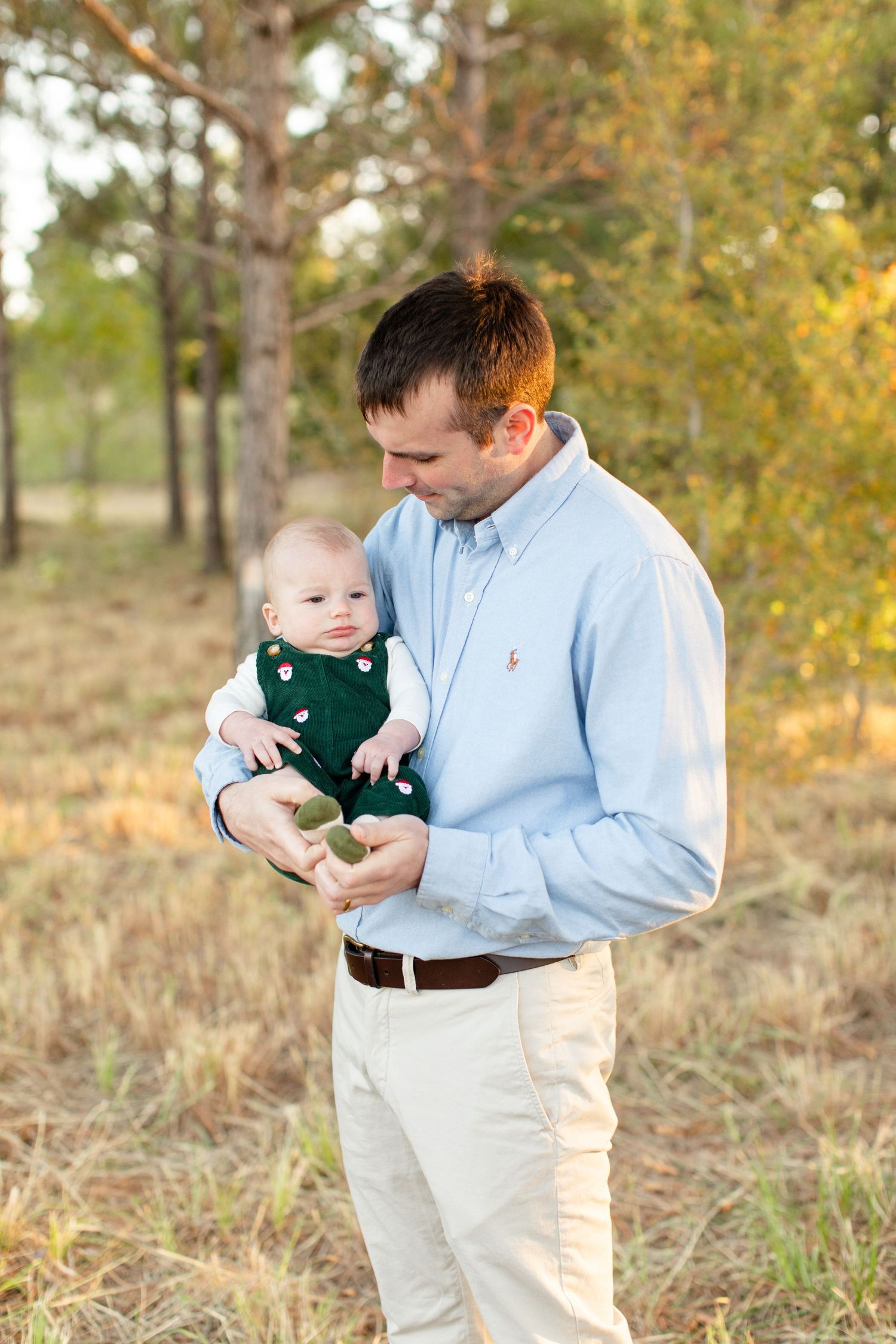 Parent in light blue shirt holding smiling baby wearing green overalls against fall foliage backdrop.