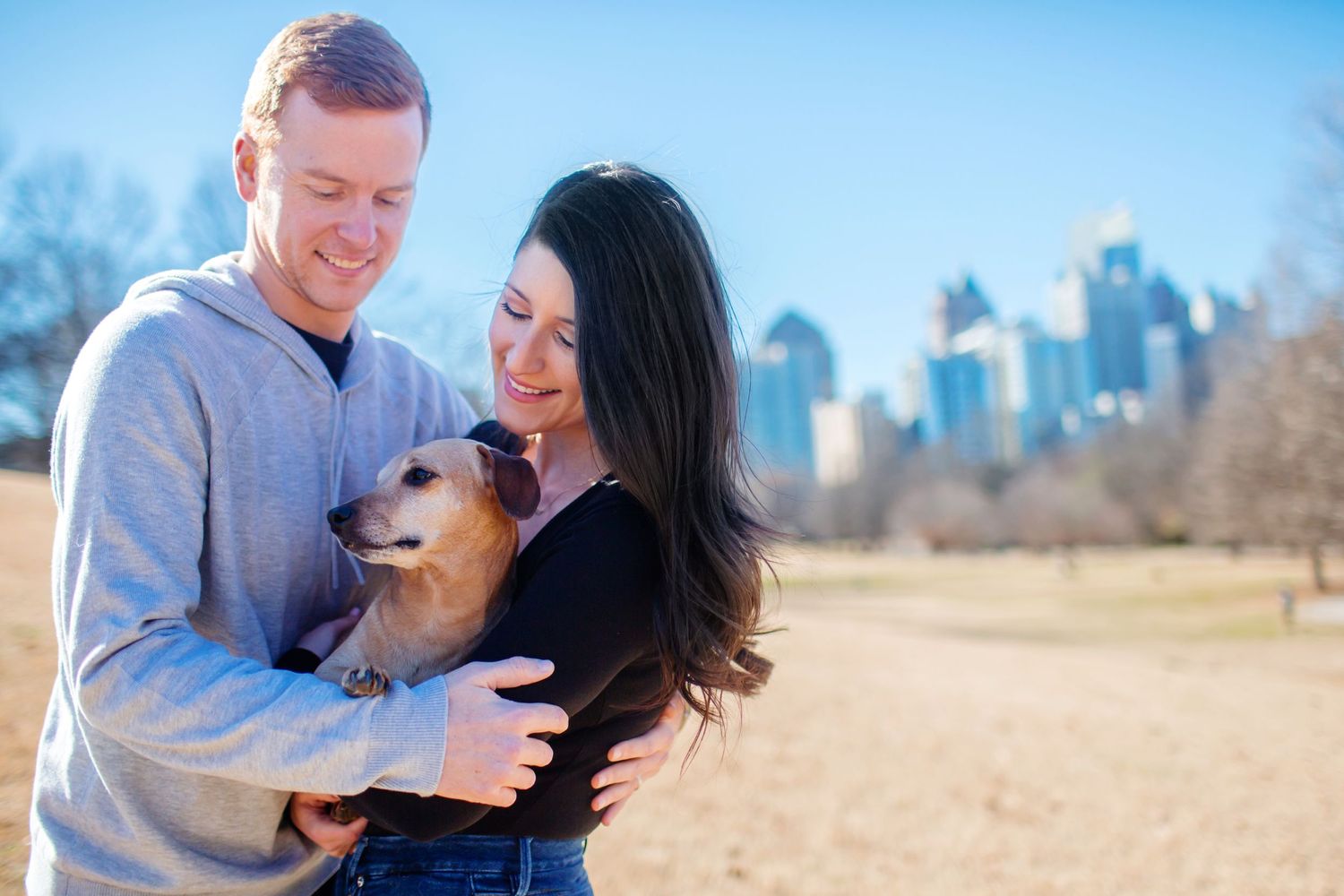 Couple embracing with their dog in front of city skyline on sunny day.