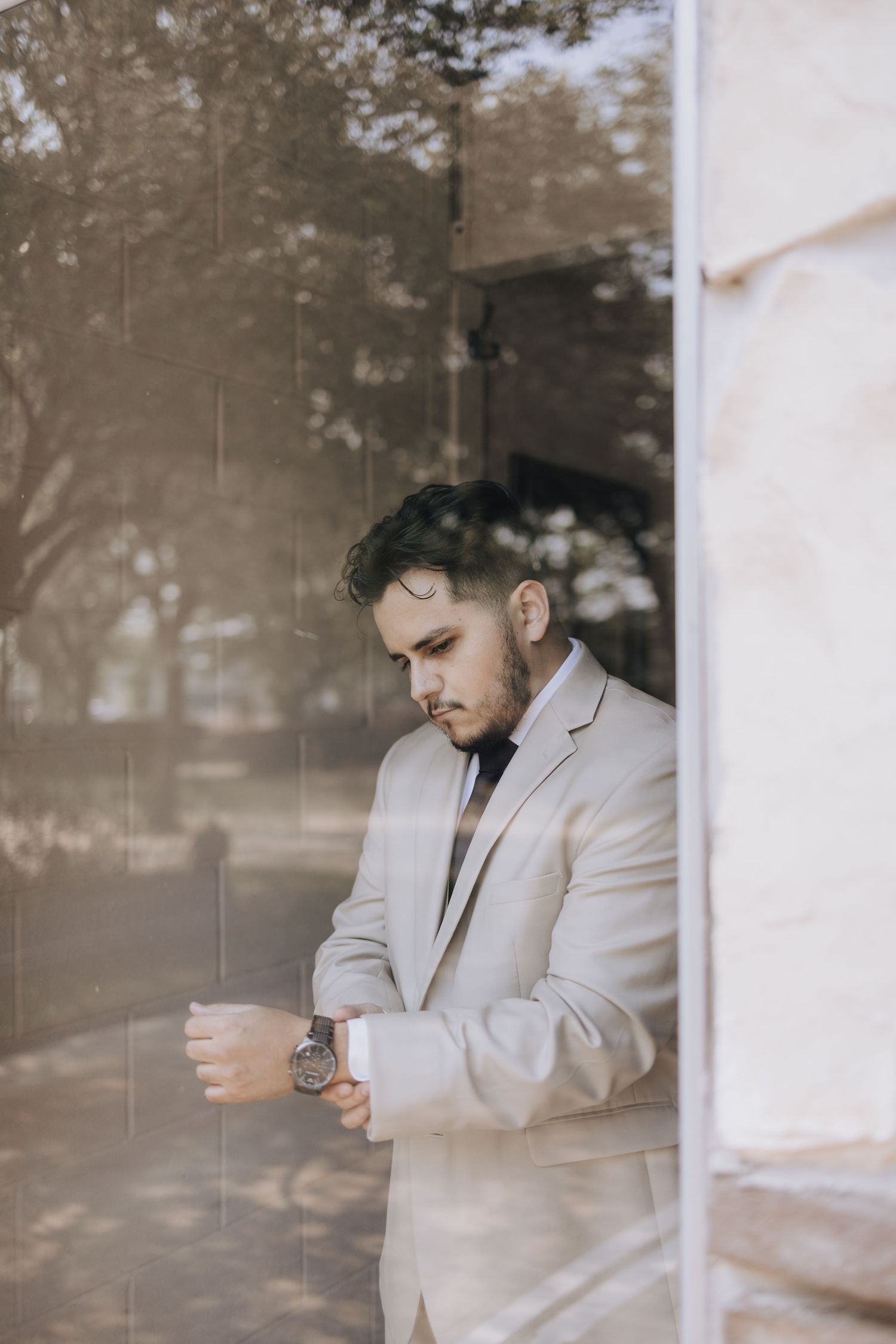 A person in a light suit adjusts their cuffs while standing by a window in contemplative pose.