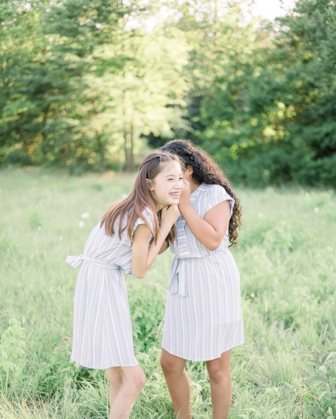Two little girls whispering in a green grassy field at golden hour.
