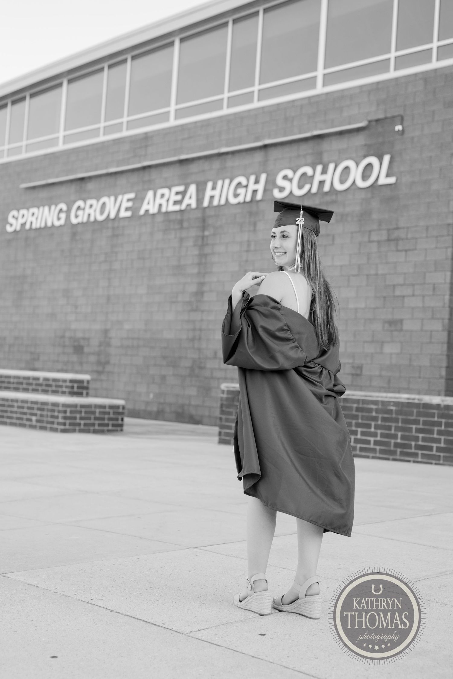Black and white portrait of graduate in cap and gown standing in front of high school building.