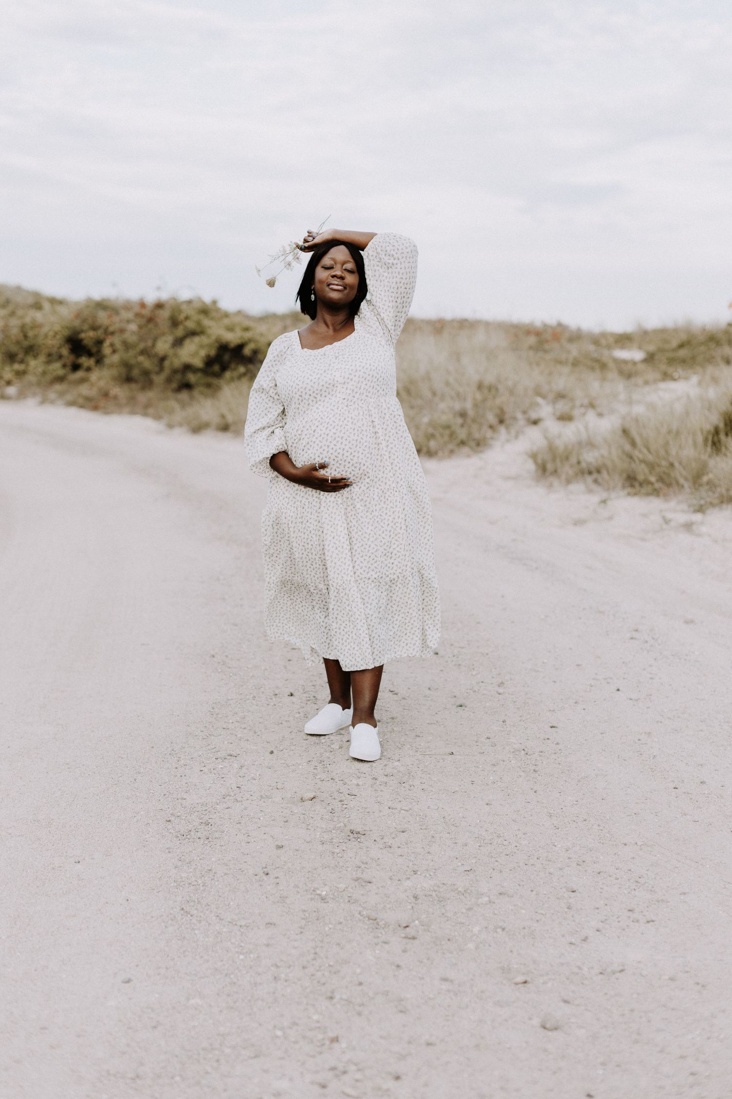 Pregnant woman in white dress walking on sandy path with hand raised above head.