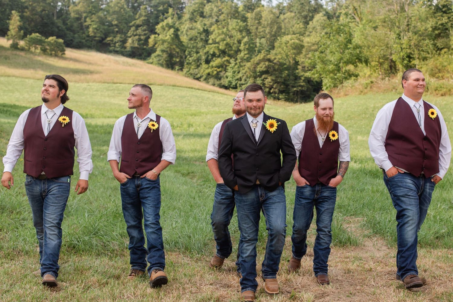 Groomsmen in burgundy vests and jeans walking across grassy field.