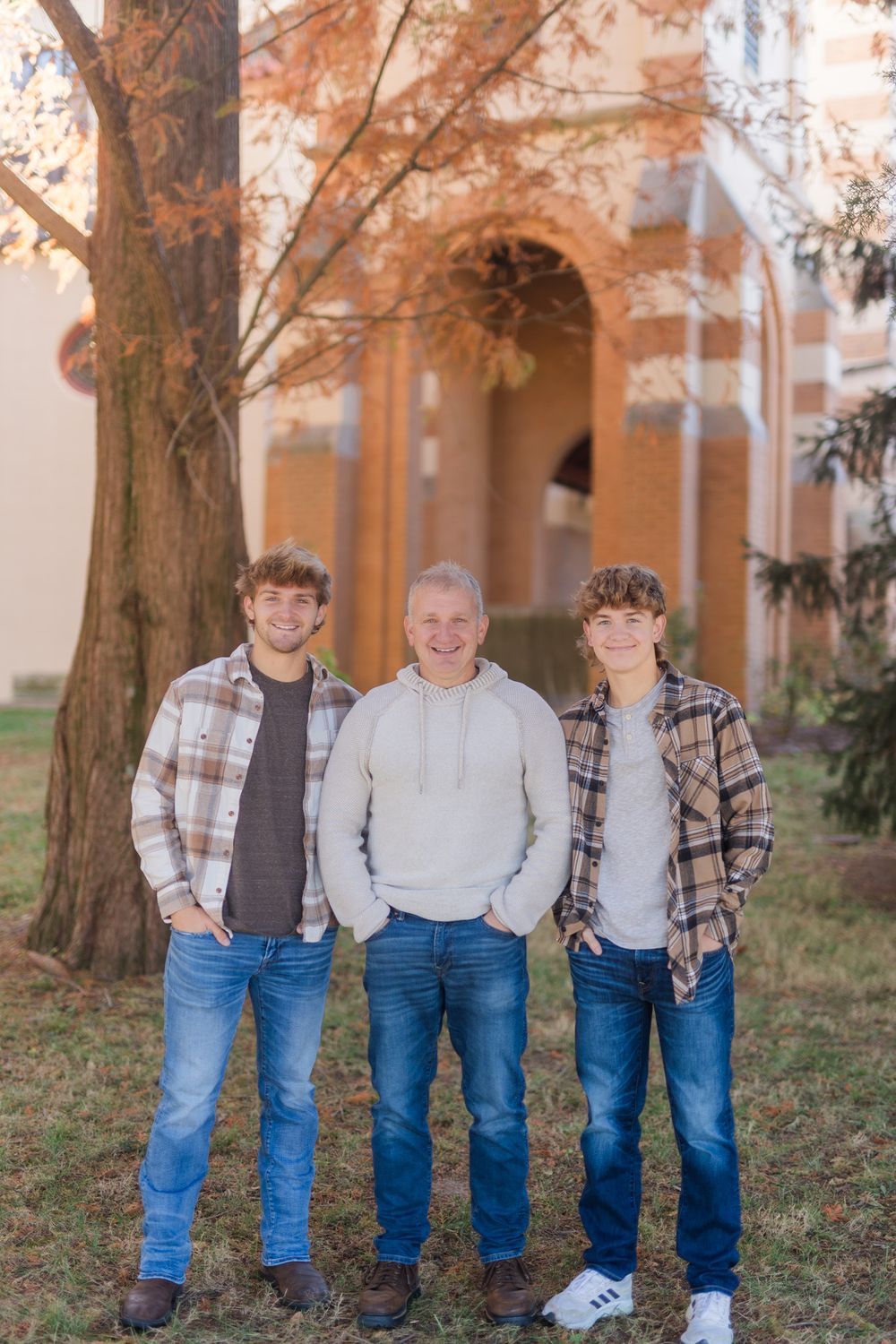 Three generations wearing casual attire pose in front of brick building on autumn day.