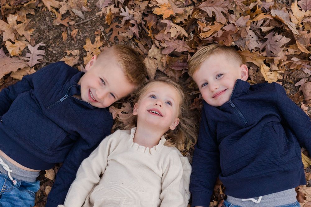 Three siblings laying together on fall leaves wearing matching navy sweaters and beige dress.