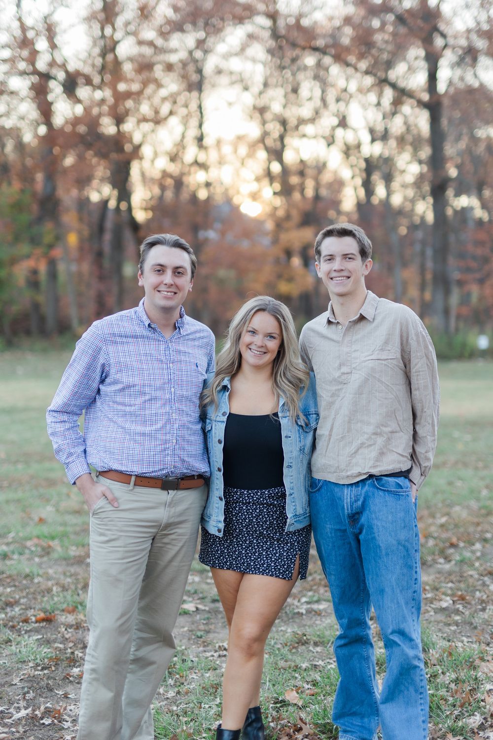 Three siblings pose together on autumn evening with sunset through trees in background.