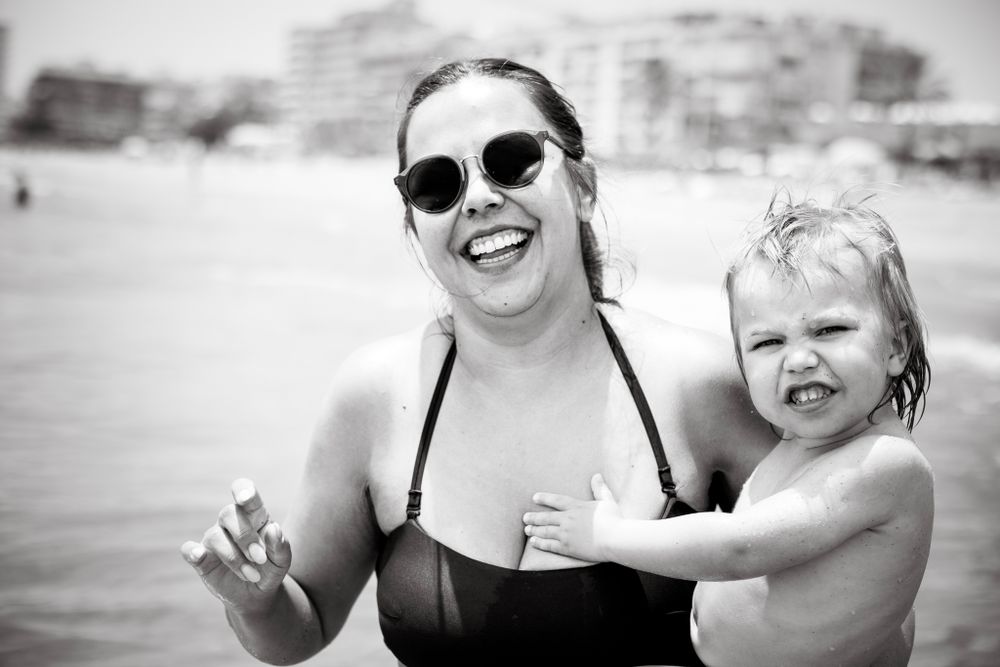 A joyful woman in sunglasses holds a squinting toddler at the beach, both smiling wide in this sunny black-and-white seaside scene.