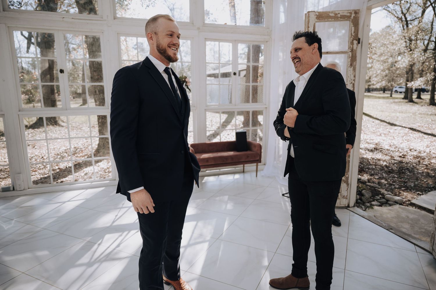Two people in dark suits having a conversation in a bright, window-filled room.
