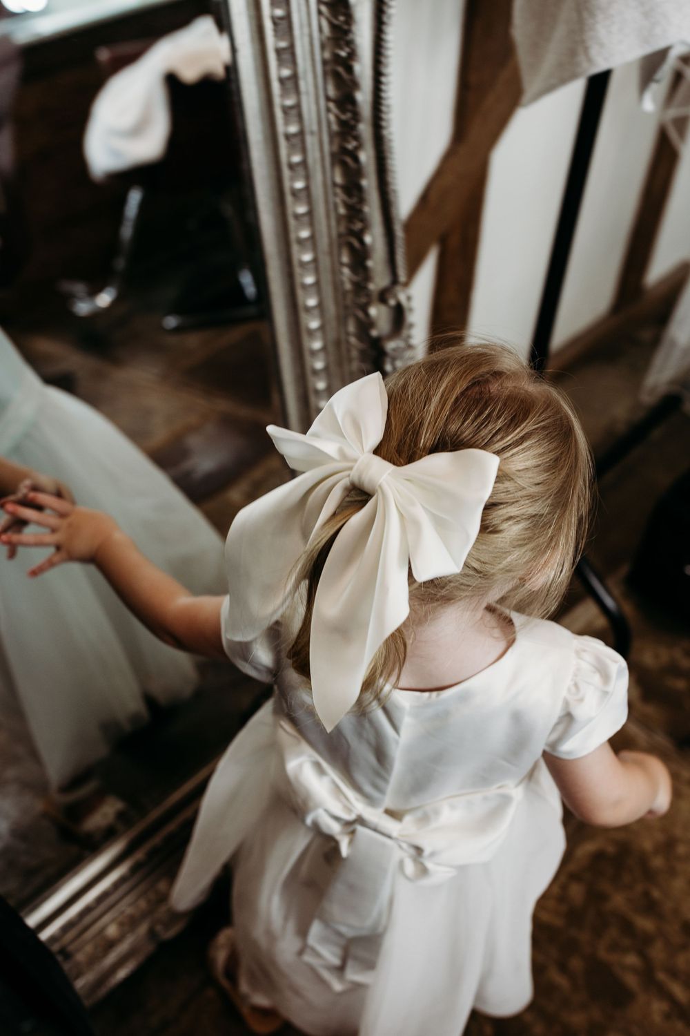A young child in a white dress with a large bow looking into an ornate mirror.